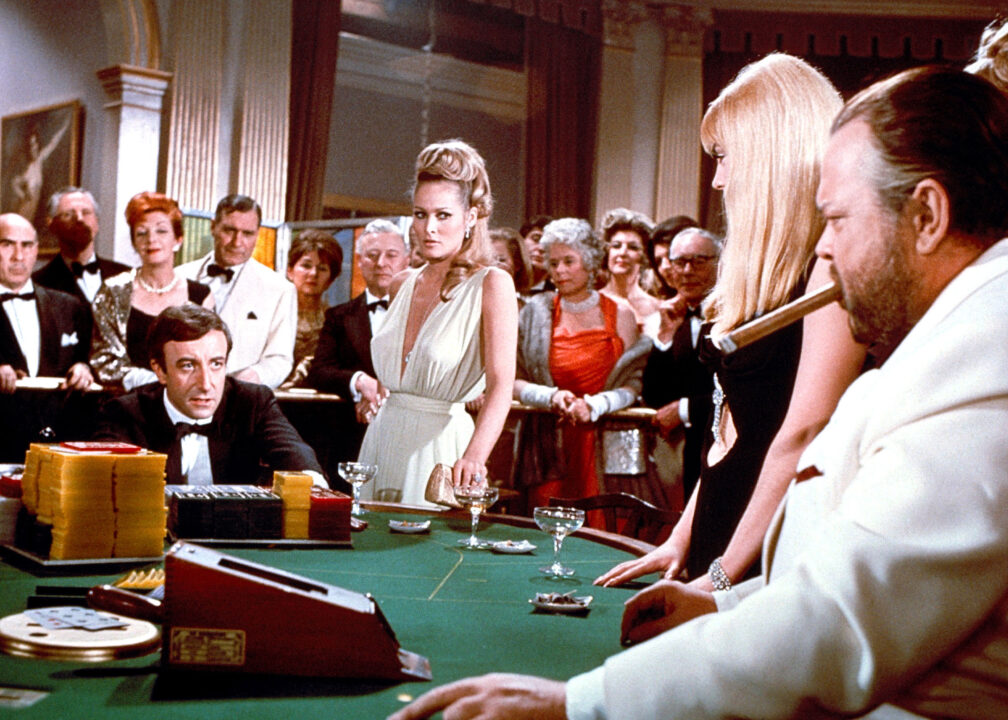 a scene from the 1967 spy spoof "Casino Royale." Seated at far left of a baccarat table, wearing a standard black-and-white tuxedo, is Peter Sellers as one of the agents dubbed "James Bond" in the film. To his immediate left, near center of the table, is Ursula Andress as Vesper Lynde (aka "James Bond"), wearing a white dress, cut quite low in the front. To far right at the table is Orson Welles as Le Chiffre; he is bearded, smoking a cigar, wearing a white tux and looking menacingly across the table at Sellers.
