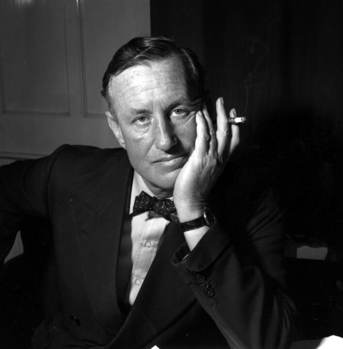 a close-up 1958 black-and-white photographic portrait of James Bond author Ian Fleming. Fleming is wearing a dark dress jacket, white dress shirt and dark bowtie with white spots. He is seated at a table, resting his cheek against his left hand (left elbow supporting on the table), and the left hand is also holding a cigarette. He has a neutral expression, not smiling.