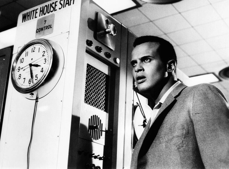  a black-and-white still from the 1959 postapocalyptic sci-fi drama "The World, the Flesh and the Devil." Star Harry Belafonte is on the right of the photo, wearing a light sport coat and white dress shirt, and looking very concerned at something (unseen to us) ahead of him and toward his right. Directly to his right, parallel to him, is some sort of computer or machine with what appears to be a speaker on it. Affixed to the front of that is an old-fashioned wall clock, whose hands are indicating it is just a little before 3:30.