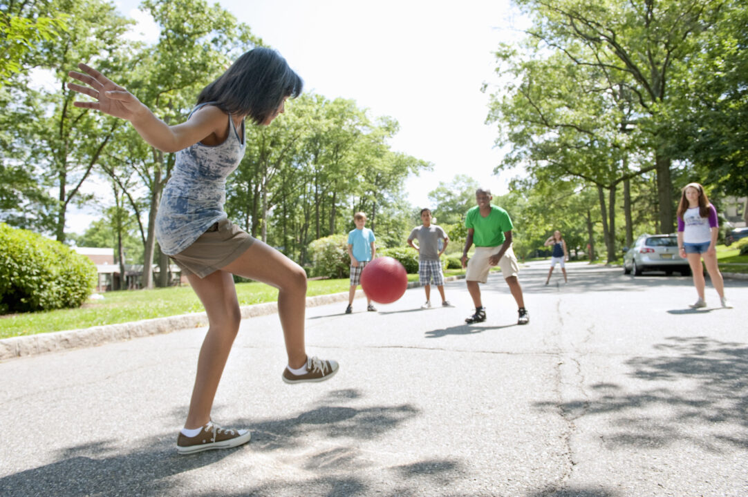 girl kicking ball to kids on opposing team, playing on a paved neighborhood street