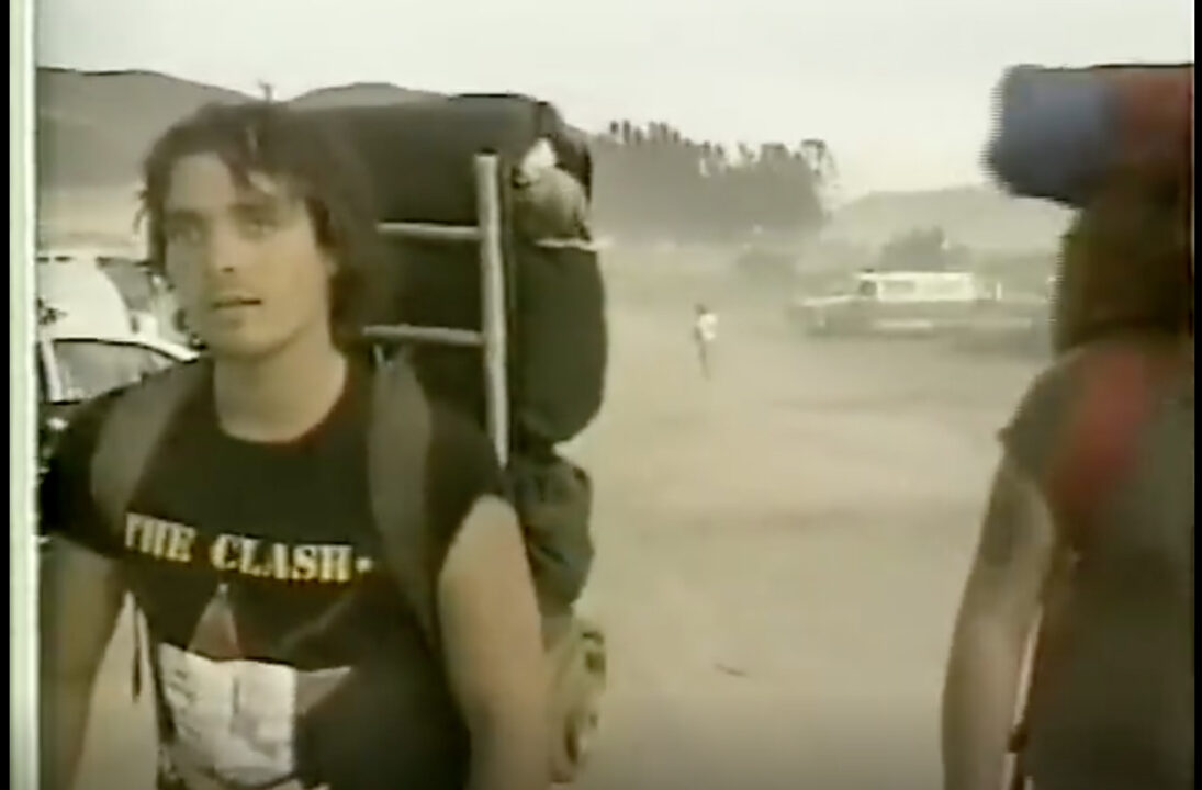 A screengrab from a YouTube video featuring film made at the 1983 US Festival, on its first day, New Wave Day. The image depicts a young man walking along the dusty road into the festival area. He has dark hair that looks sweaty, and if he has been walking a while to get in, and his eyes are somewhat dazed as he walks past the camera. He is wearing a large, camping/hiking-style backpack and a black T-shirt with an image and logo for the band The Clash, who were one of the groups performing that day.