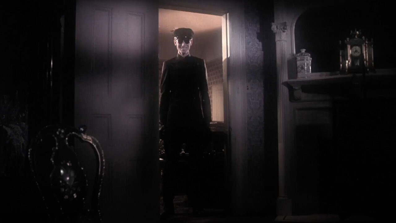 A scene from "Burnt Offerings" (1976), with Anthony James as the sinister chauffeur, standing in a doorway and grinning malevolently into the room at Oliver Reed and Bette Davis' characters (unseen in the photo). It is a wide shot, so we see the chauffeur completely standing in the doorway, dressed in an old-time chauffeur's cap and suit, wearing sunglasses.