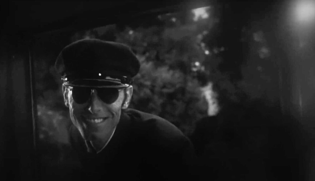 Anthony James as the chauffeur/hearse driver in the 1976 horror movie "Burnt Offerings." The image is in black and white, in a nightmare scene, as the chauffeur -- wearing an old-time cap and suit, and aviator sunglasses -- suddenly flashes a sinister-looking smile through the window after slamming the car door behind young Ben (not seen in the photo) at his mother's funeral.