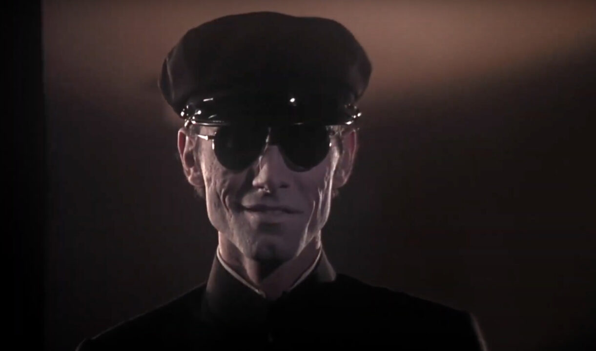a closeup shot of Anthony James as the chauffeur in the 1976 horror movie "Burnt Offerings." He is just starting to crack the malevolent smile he flashes throughout the movie. He is wearing an old-time chauffeur's cap and sunglasses in this nighttime image.