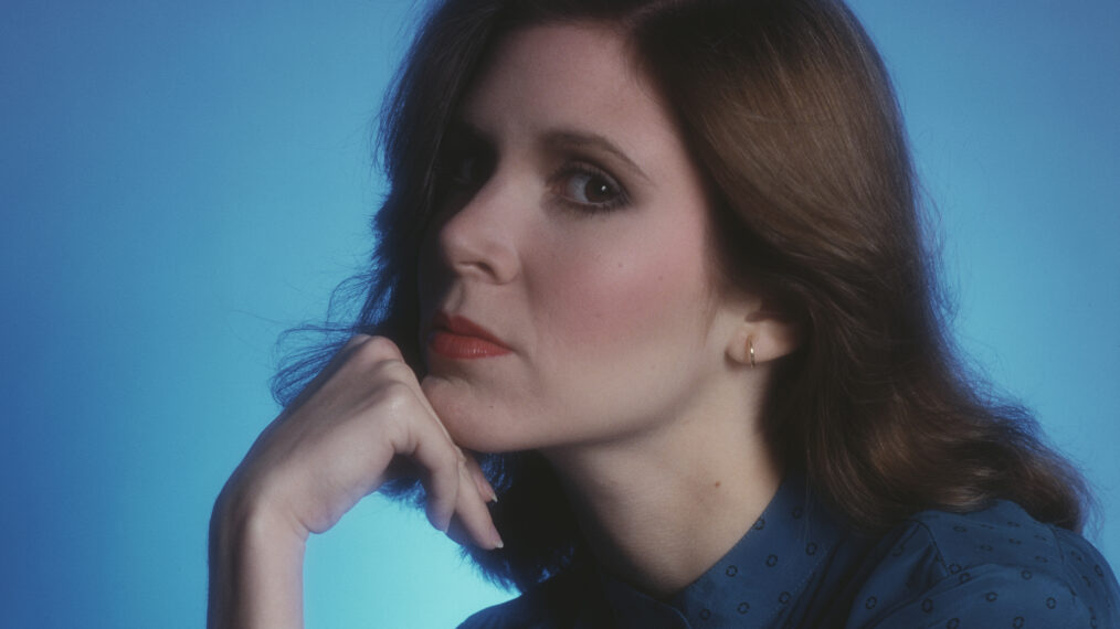 a 1980 portrait photo of actress Carrie Fisher. She is seated in profile, with her left side facing viewer, with her right hand on her cheek and against a light blue background. She has longer, shoulder-length brown hair.