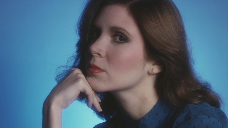a 1980 portrait photo of actress Carrie Fisher. She is seated in profile, with her left side facing viewer, with her right hand on her cheek and against a light blue background. She has longer, shoulder-length brown hair.