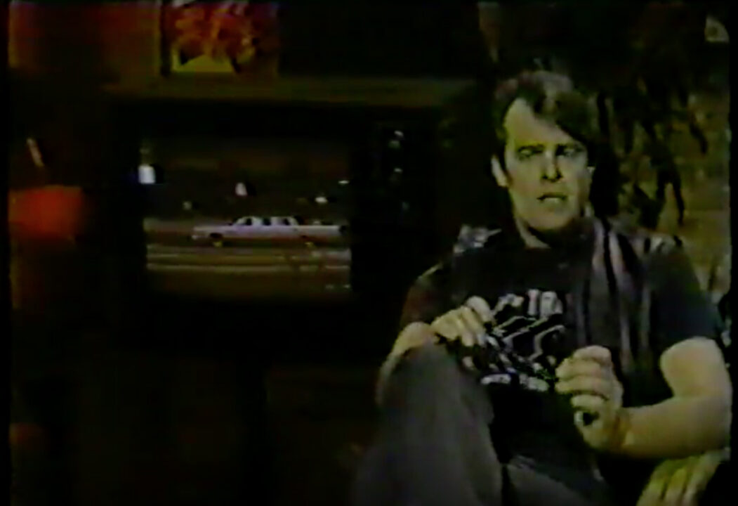 Dan Aykroyd as MTV guest VJ in May 1983. He is seated, holding his glasses in both hands as he discusses MTV's contest related to his film "Doctor Detroit." The grand prize is the stretch limo seen in the movie, which is depicted on a TV screen behind Aykroyd and to his right.