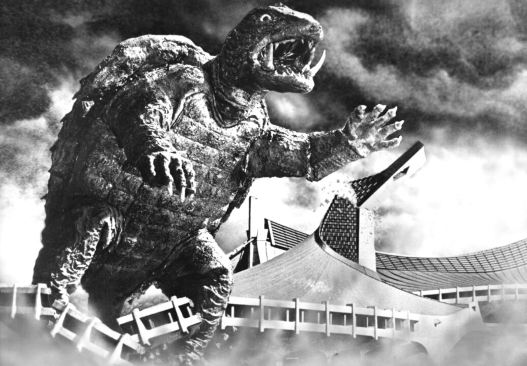 an image from the 1966 Japanese kaiju movie "Gamera the Invincible." It is a black and white image depicting Gamera, a giant prehistoric, bipedal turtle, stomping over a building as he roars and exposes his two large fangs growing upward from the bottom of his jaw.