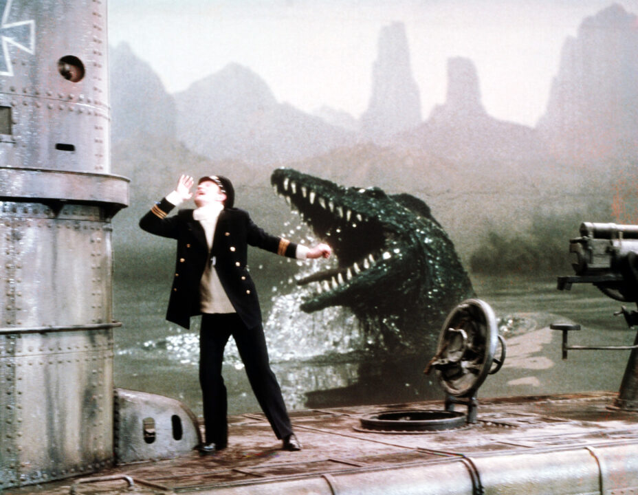  a still from the 1974 movie "The Land That Time Forgot." It depicts a World War I German U-boat which has surfaced in the waters of a previously hidden prehistoric world. A German officer is standing on top of the boat and beginning to run as, behind him, an enormous, green, crocodile-like Mososaur comes out of the water and lunges at him with its jaws wide open, exposing lots of sharp white teeth.