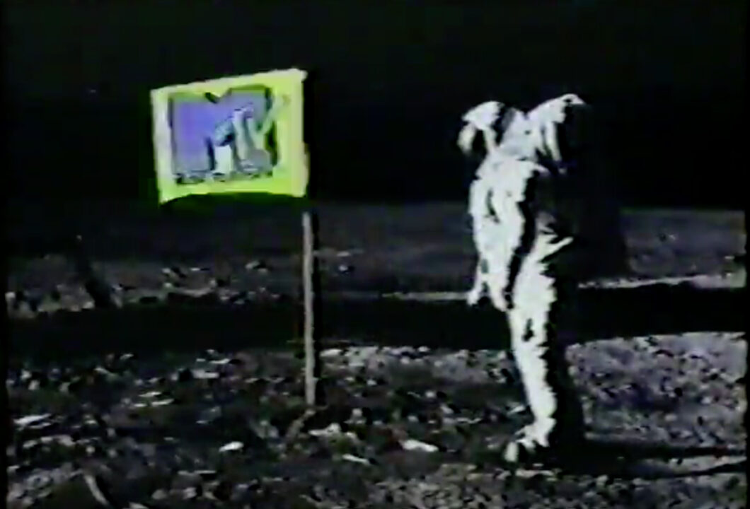 a shot from the MTV "Moon Man" network ID spot from May 1983. It depicts an astronaut on the moon on the right side of the photo, looking toward the left side, on which a flag is planted on the moon. The flag features MTV's logo.