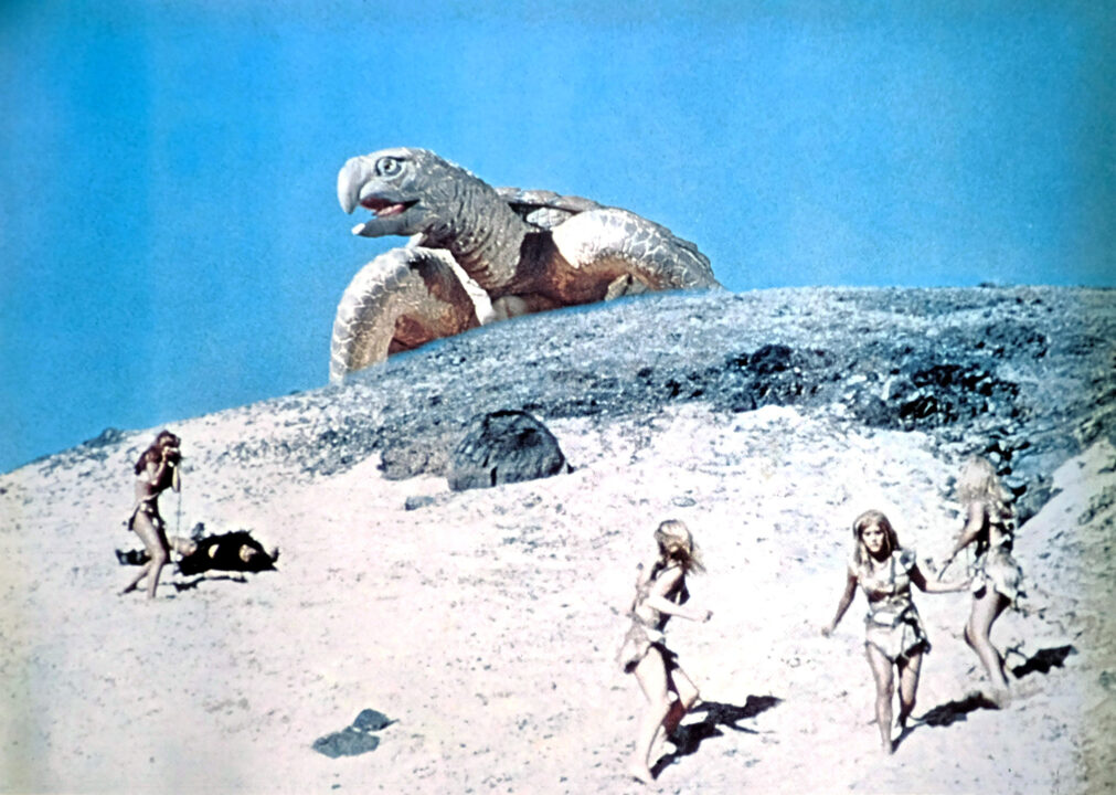 a scene from the 1966 prehistoric-set fantasy movie "One Million Years B.C." A group of the film's cavepeople characters are at the bottom of a sand dune on a beach, looking up at the top of the dune, over which, against a blue sky in the background, is an approaching gigantic Archelon sea turtle that has just come out of the ocean, in a special effect created by Ray Harryhausen.