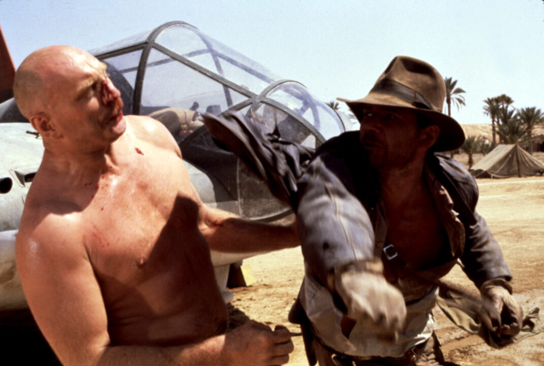 Pat Roach (left) as a Nazi mechanic immediately after being punched by Indiana Jones (Harrison Ford, on the right) in a fight scene from "Raiders of the Lost Ark." The mechanic is large, bald, shirtless, sports a mustache and looks imposing. His nose is bleeding after this and other previous punches from Jones, and he is falling back a bit. Jones' arm and fist are in the follow-through after punching the Nazi, and he has a determined look on his face, which is also bloodied from taking his own punches. He is wearing his traditional brown leather jacket and brown fedora with black band. They are in closeup, fighting in front of a "flying wing" plane, the cockpit of which can be seen immediately behind them.