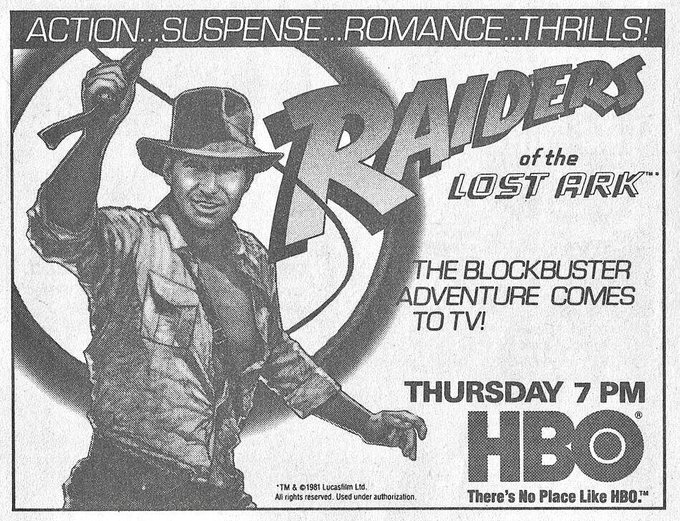 a black-and-white print ad for the television premiere of "Raiders of the Lost Ark" in 1984. The ad is in a small square area. Taking up the left of the ad is an image from the movie's theatrical poster, with a drawing of Harrison Ford as Indiana Jones in the process of cracking his bullwhip. Immediately to his upper right is the movie's title treatment, with "Raiders of the Lost Ark" presented in the old-style adventure film font. Below that reads the message: "The blockbuster adventure comes to TV!" Below that says "Thursday 7pm" right above the HBO logo (with the slogan "There's no place like HBO" right below that. Running across the top of the ad in a thin strip reads the hype line "Action ... Suspense ... Romance ... Thrills!"