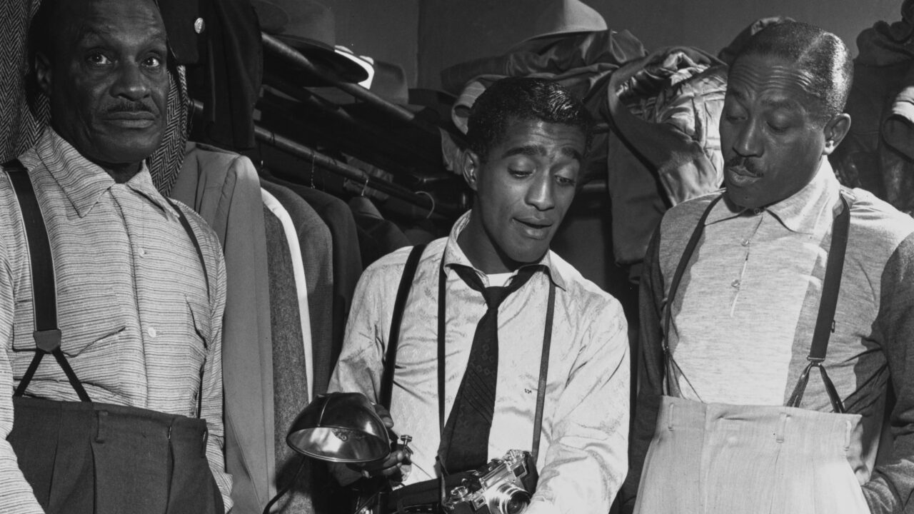 American singer and actor Sammy Davis Jr. (1925 - 1990) (center) shows his photography equipment to his father, Sammy Davis Sr. (left), and his 'uncle' Will Mastin in a dressing room, 1953. The three men had comprised the Vaudeville act, The Will Mastin Trio, following World War II. 