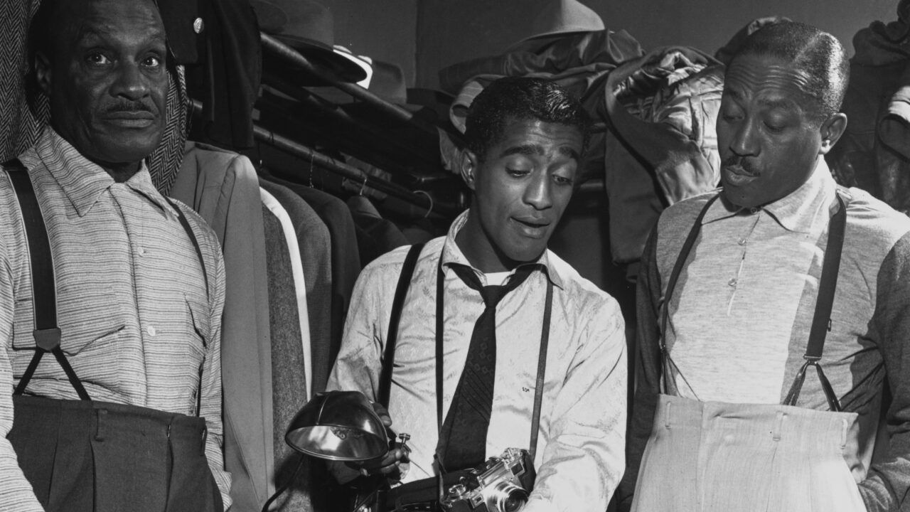American singer and actor Sammy Davis Jr. (1925 - 1990) (center) shows his photography equipment to his father, Sammy Davis Sr. (left), and his 'uncle' Will Mastin in a dressing room, 1953. The three men had comprised the Vaudeville act, The Will Mastin Trio, following World War II.