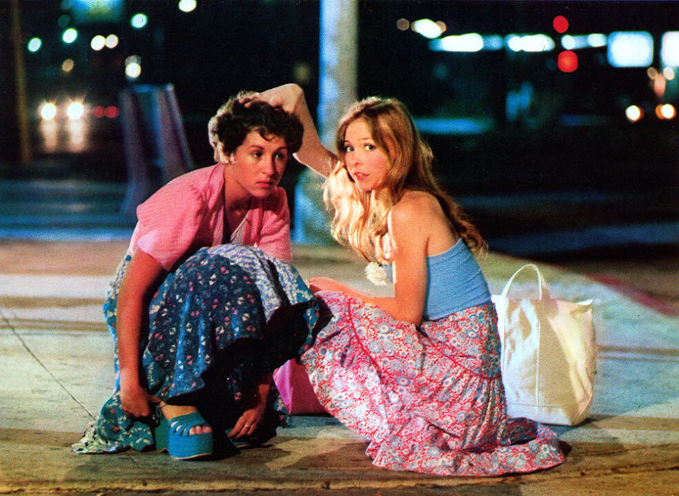 shot from the 1978 movie "Thank God It's Friday" depicting the characters Frannie (Valerie Landsburg) and Jeannie (Terri Nunn). They are both crouching on a sidewalk, Frannie on the left, Jeannie on the right, looking toward the camera and at something unseen to the viewer of the photo. 