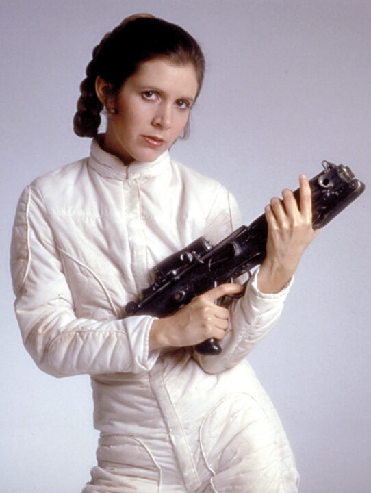 Carrie Fisher in a promotional photo from "The Empire Strikes Back" (1980). She is dressed in the all-white snowgear she wears in the film's opening scenes on the icy planet Hoth. She is holding a long laser blaster in both hands, with a determined look on her face.