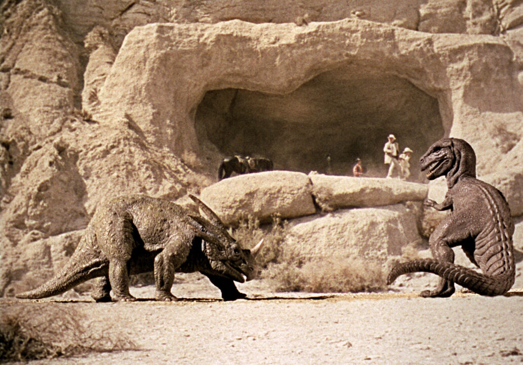 an image from the 1969 movie "The Valley of Gwangi." It is a stop-motion special effects shot of two dinosaurs -- a horned Styracosaurus on the left, a carnivorous, T. rex-like Allosaurus on the right, about to fight in front of a desert cave, where some human bystanders are looking on.