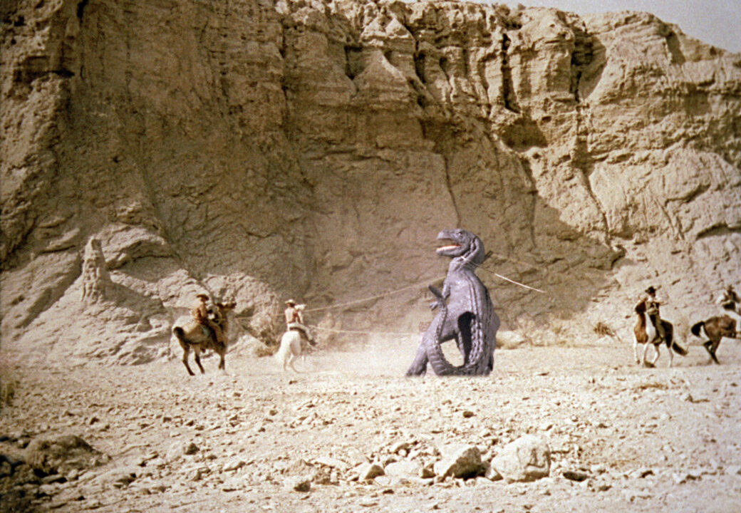 still from the 1969 movie "The Valley of Gwangi." it is a stop-motion special effects shop detailing a group of cowboys on horseback in a mountainous desert environment who are surrounding an Allosaurus dinosaur and roping it with their lassos.