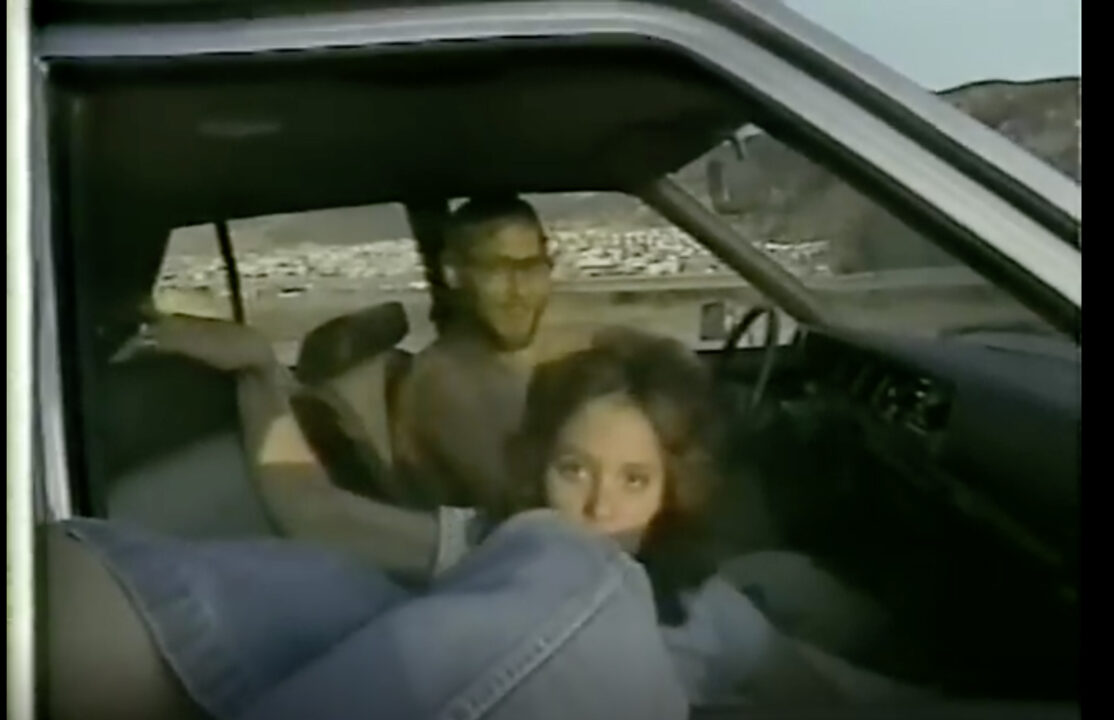 screengrab from a YouTube video featuring film footage from the 1983 US Festival. The image is a closeup of a car, taken from just outside the passenger side. The car is stopped and waiting in line to get into the festival. A shirtless young man with glasses and a mustache is behind the wheel. In the passenger seat, laying with her feet and legs dangling out the window and lifting her head so it can just be seen, is the young woman accompanying him.