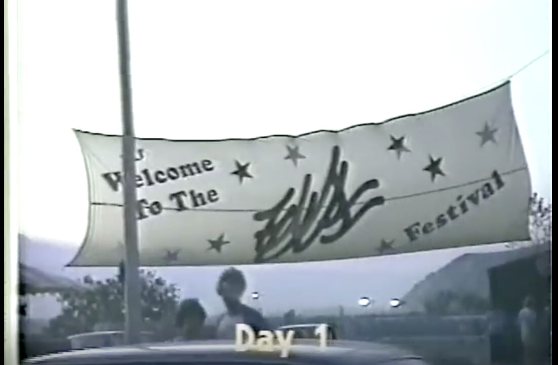 A banner across the road on the way into the 1983 US music festival. It is a white banner with a few black stars on it, and in dark lettering reads "Welcome to the US Festival." It is in closeup, but you can tell there are cars lined up beneath it entering the festival, and a bit of the crowd of people can be standing to the side. At the bottom of the image reads a graphic superimposed upon the video of the event from which this screengrab was taken that says, in yellow lettering: "Day 1."