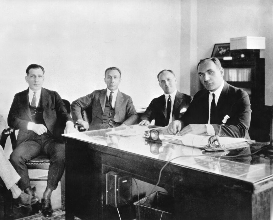 1926 black-and-white photo of the Warner brothers, the siblings who founded Warner Bros. Seated from left to right are Sam, Harry, Jack and Albert. Albert is seated at a desk, with the other three brothers along the side of the desk to his right.