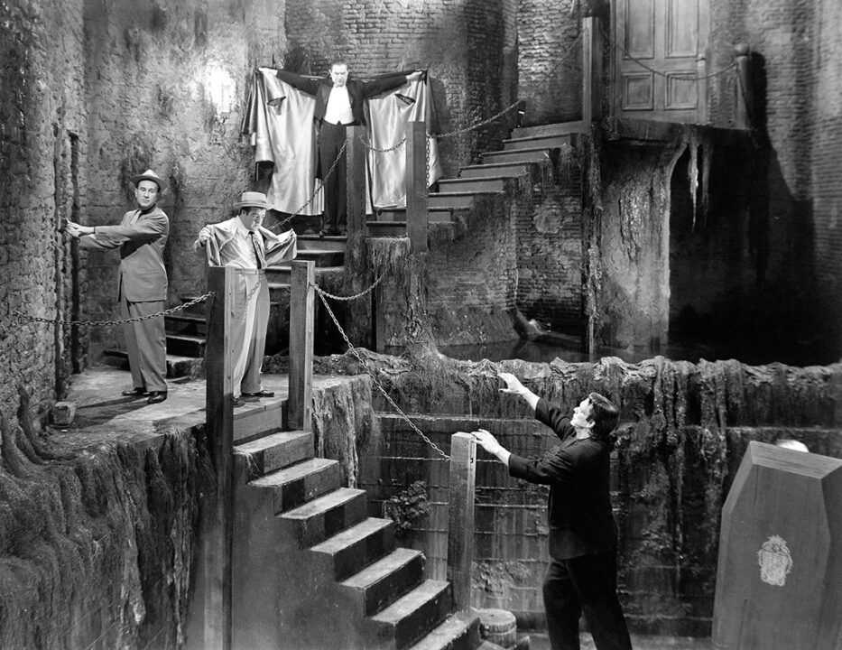 black and white shot from the 1948 horror comedy "Abbott and Costello Meet Frankenstein." The scene is in a spooky old brick building, with tiered staircases leading up toward the left, then angling up to the right. Coming down the stairs on the right is Bela Lugosi as Count Dracula, dressed in his black cape, which is raised as Dracula has both arms outstretched while he comes down the stairs and approaches Bud Abbott, on the left, who is trying to open a door, and Lou Costello, just to his right. Both men are dressed in suits and hats of the time (1940s). Lou is looking down the flight of stairs heading up to the left, at the top of which he is standing. He his his suit jacket outstretched much the same way Dracula has his cape outstretched. Lou is looking toward the bottom of the stairs, because heading toward them and about to climb up them is the Frankenstein monster (Glenn Strange) with outstretched arms.