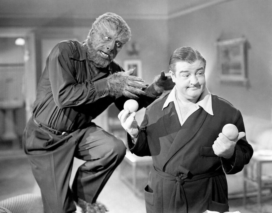 black and white shot from the 1948 horror comedy "Abbott and Costello Meet Frankenstein." In the left of the close-up image is Lon Chaney Jr. in costume as the Wolf Man, who is balanced on his right leg with his left leg in the air, bent at the knee, looking to be about to pounce, as his hands/claws are in a position looking about to clasp around the next of an oblivious Wilber Gray (Lou Costello) right next to him, on the photos' right. Wilbur is holding up an egg in each of his hands and smiling as he looks at them, unaware of the Wolf Man right next to him.