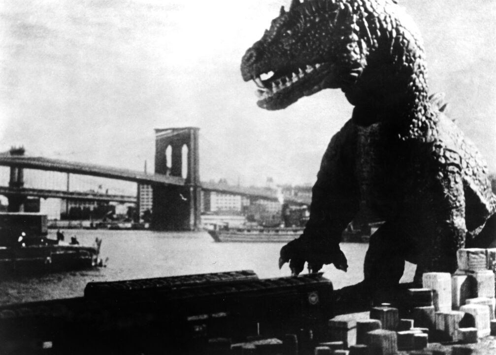 black-and-white still image from the 1953 monster movie "The Beast From 20,000 Fathoms." It depicts the dinosaur-like "Rhedosaurus" coming out of the harbor into New York City to begin its rampage. In the backdrop is an image of a bridge and the city skyline. The creature is scaled, with spikes on its head and baring its teeth as it roars.