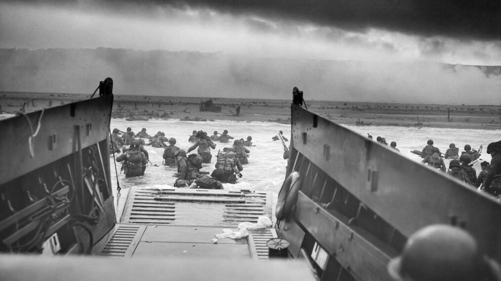 Digitally restored, vintage black-and-white World War II photo of American troops wading ashore on Omaha Beach during the D-Day invasion on June 6, 1944. The photo is taken from the perspective of the photographer standing in a landing craft behind the soldiers who are leaving the lander and heading through the surf toward the beach.