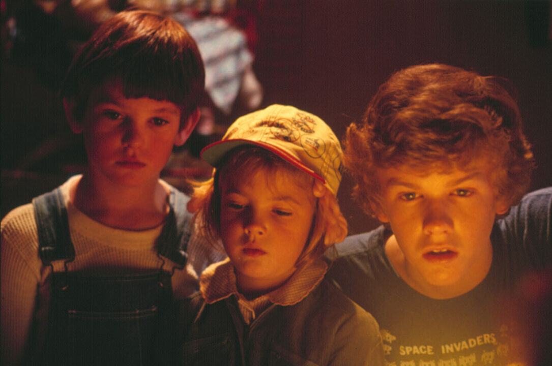 a still image from the 1982 movie "E.T. the Extra-Terrestrial" Left to right are Henry Thomas, Drew Barrymore and Robert MacNaughton, in closeup, staring in awe at E.T. (not seen in the image).
