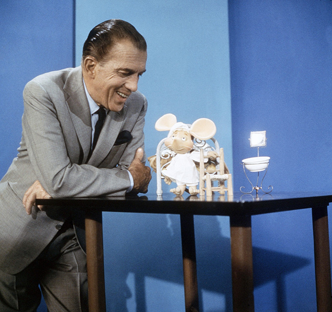 an image from an early 1960s episode of "The Ed Sullivan Show. It is a color photo of Ed, leaning on a table with crossed arms and smiling as he looks down at Top Gigio, his "Little Italian Mouse" puppet occasional sidekick