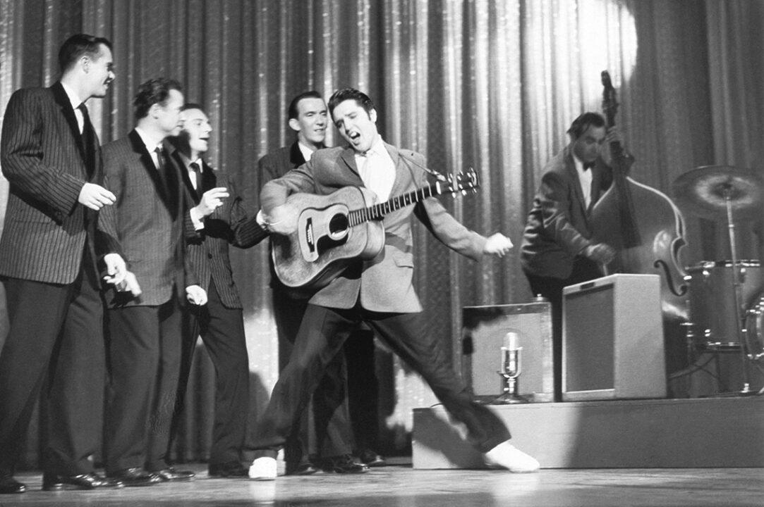 black-and-white photo of Elvis Presley's second appearance on "The Ed Sullivan Show," Oct. 28, 1956. Elvis is holding a guitar in his right hand while gyrating and extending his left arm while singing and dancing onstage, as his backup singers, the Jordanaires, accompany him just behind.
