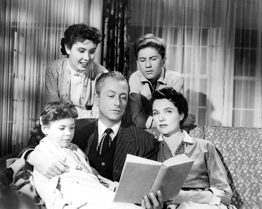 a black-and-white cast shot from the 1954-60 sitcom "Father Knows Best." Robert Young, as family patriarch Jim Anderson, is sitting on a couch, holding an open book in one hand and appearing to be reading from it aloud. Sitting on his lap, on his right, is Lauren Chapin as his youngest daughter, who is listening to him read and also looking at the book. Close to him, and sitting on the couch to his left, is Jane Wyatt as his wife, Margaret. Standing right behind him and looking down at the book and listening are (on his right) Elinor Donahue as oldest daughter Betty, and (on his left) Billy Gray as son Jim Jr.