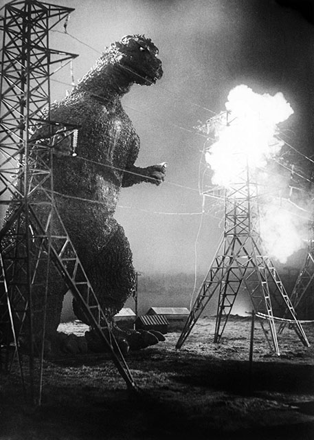 a black-and-white image from the 1954 Japanese movie "Gojira" ("Godzilla"). Godzilla is on the left left of the photo, heading toward the right and about to knock down power lines stretched across his path as he walks between two electrical towers. Fire blazes in the background from other devastation.