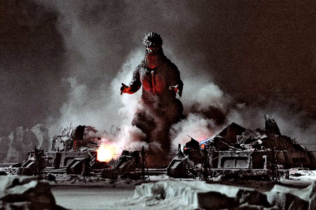 a scene from the 2004 movie "Godzilla: Final Wars." It depicts the dinosaur-like monster Godzilla a bit far off in the background, approaching toward the front of the photo, walking through a devastated city still smoldering and smoking in the night.