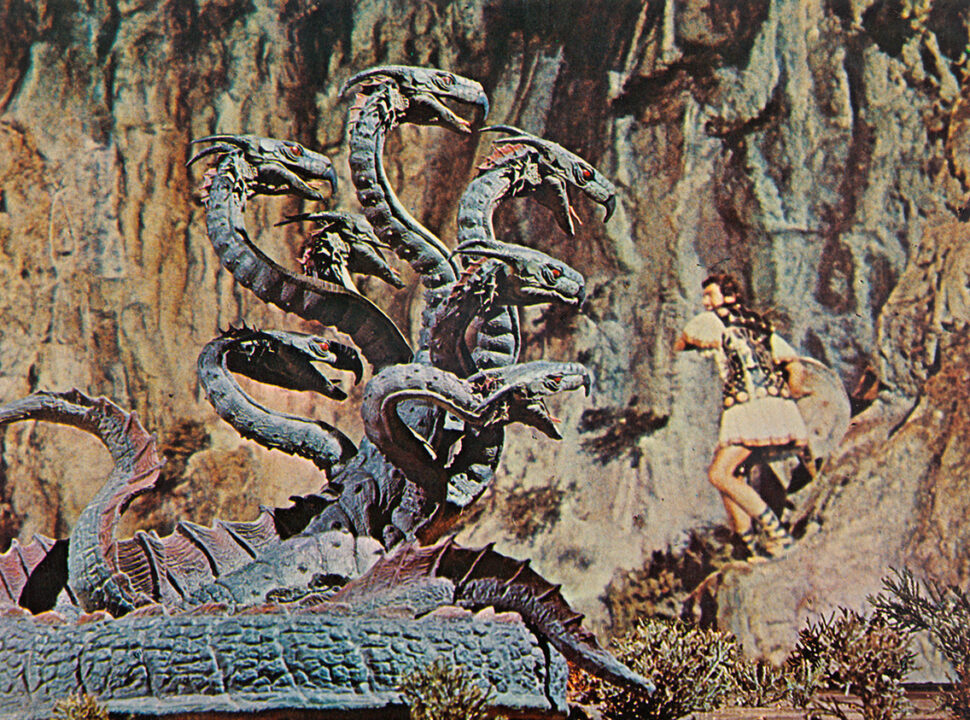 shot from the 1963 movie "Jason and the Argonauts," depicting Jason battling the Hydra, a creature with seven snake-like heads. Jason is standing atop a rock outcropping to the right of the photo, with the Hydra approaching near him from the left, its serpentine heads all gazing on him as he is about to swing his sword at it.