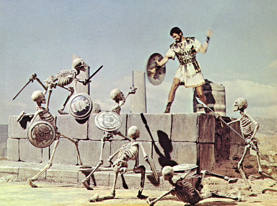 scene from the 1963 movie "Jason and the Argonauts." It depicts the fight between Jason and a skeleton army created by special effects. The bearded Jason is standing atop a small portion of ruins, wielding a shield in his right hand and a sword, raised above his head about to strike, in his left. He is looking down at six sword-and-shield-carrying skeletons who are converging to attack him.