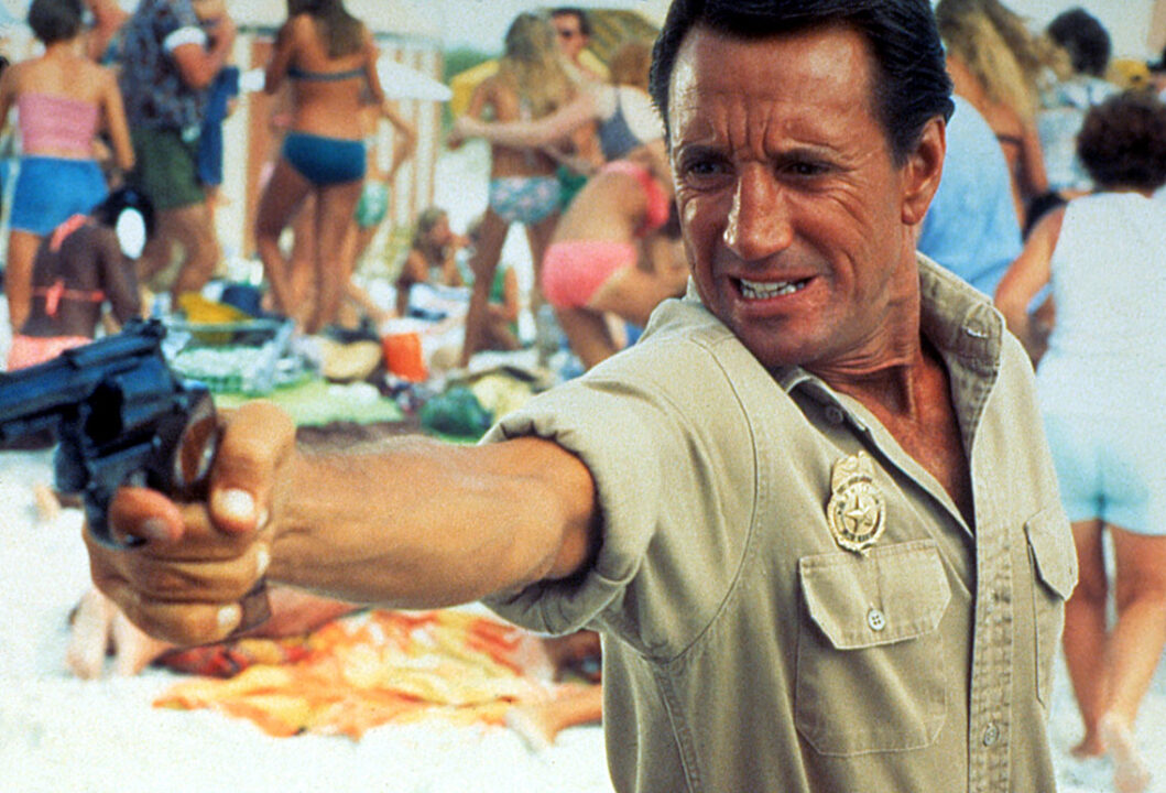 photo still from the 1978 movie "Jaws 2." It is a closeup of Roy Scheider as Chief Brody, who is on a beach and angrily pointing his gun, with an outstretched arm, presumably aiming toward the water and the killer shark.