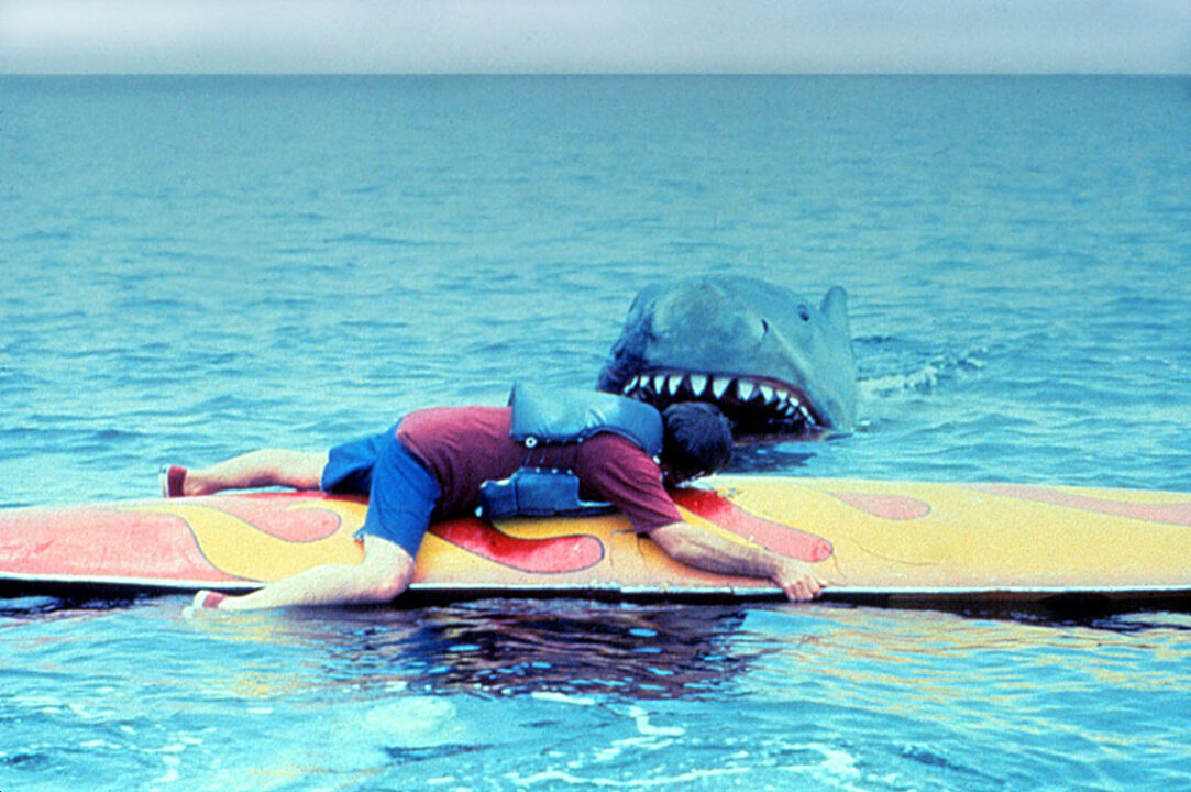 a photo still from the 1978 movie "Jaws 2." It depicts a man wearing a life preserver and clinging to a surfboard, in the foreground of the photo, looking away toward a shark that is approaching. The shark's head is a bit out of the water, revealing its eyes and teeth in an open mouth that is about to attack the man.