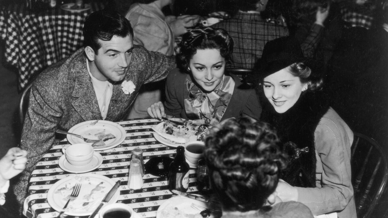 circa 1940: Olivia De Havilland leans in to hear a conversation while lunching with her sister Joan Fontaine and actor John Payne. 