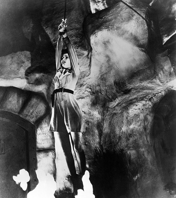 black and white still from the 1942 movie serial "Perils of Nyoka." Kay Aldridge, as Nyoka, is in one of her character's frequent cliffhanger situations. She is hanging by her rope-bound hands over a pit in which flames are burning, and has a frightened look on her face.