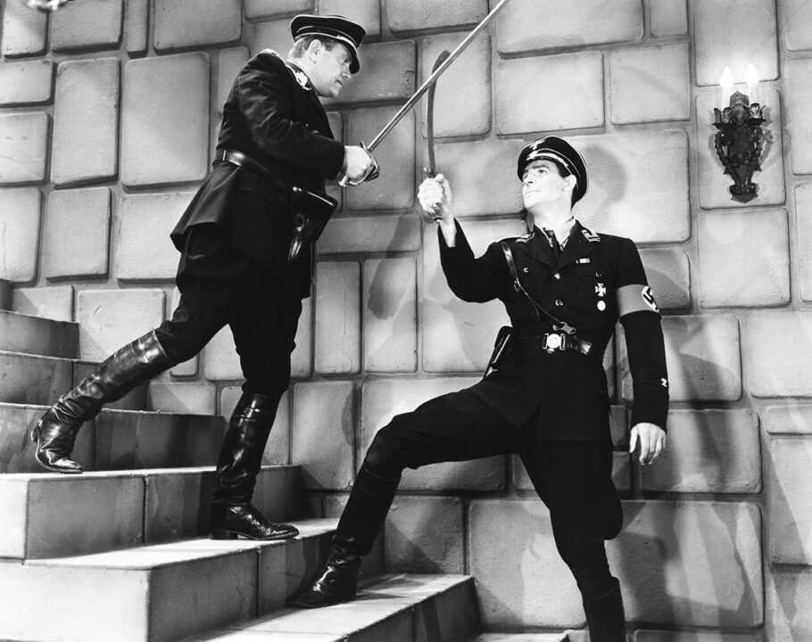 black and white still from the 1943 movie serial "Secret Service in Darkest Africa." Two men are dueling with swords on a stone staircase against a stone wall. The man at the bottom, facing up at his opponent a couple of steps above him, is an undercover agent dressed in a Nazi officer uniform. The man he is dueling is an actual Nazi foe, also wearing an officer's uniform.