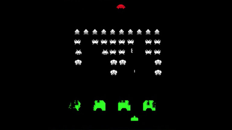 a screen shot from the 1978 arcade video game Space Invaders, depicting the player's green ship along the bottom, behind four green shields, firing up at rows of white-colored invaders getting increasingly lower and firing back. A red UFO is moving across the top of the screen.