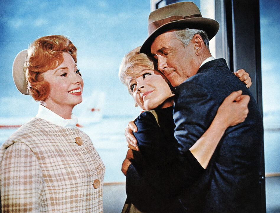 a color still from the 1963 movie "Take Her, She's Mine." left to right are Audrey Meadows, Sandra Dee and James Stewart. Stewart, wearing a fedora hat and coat, is hugging Dee, who plays his daughter, as Meadows looks at them happily, wearing a pillbox hat.
