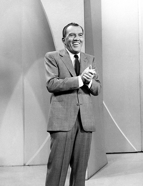 black and white shot of Ed Sullivan onstage to introduce "The Ed Sullivan Show." He is smiling and has his hands clasped together.