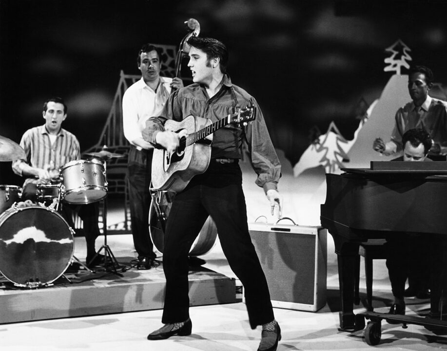 black-and-white photo of Elvis Presley making his first appearance on "The Ed Sullivan Show" on Sept. 9, 1956. He is onstage, in front of a piano and his backup band, holding a guitar in one hand.