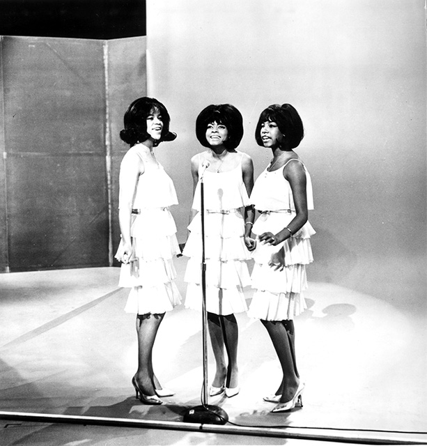 black and white photo of The Supremes making their first appearance on "The Ed Sullivan Show" in December 1964. The three women are wearing white dresses and standing on the stage. Left to right are Florence Ballard, Diana Ross and Mary Wilson.