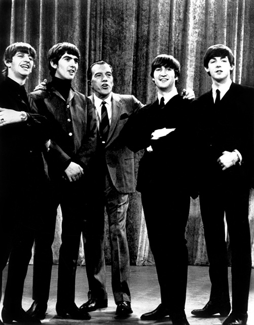 black-and-white photo of the Beatles with Ed Sullivan during their first appearance on his show on Feb. 9, 1964. Sullivan is in the center of the stage between George Harrison and Paul McCartney to his right, and John Lennon and Ringo Starr to his left.