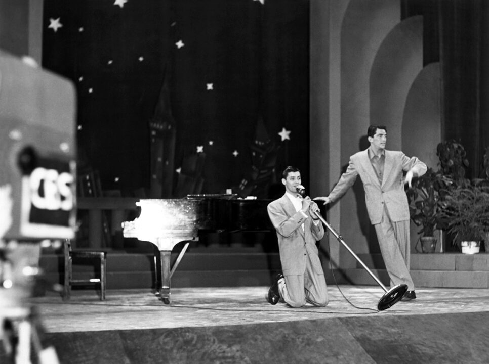 black-and-white image from the first episode of "The Ed Sullivan Show" (known as the time as "Toast of the Town" on June 20, 1948, Pictured is the comedy team of Dean Martin and Jerry Lewis performing onstage. Lewis is kneeling and holding a long microphone stand and mic right up to his mouth, with Martin standing at an angle to Lewis' left, resting his right hand on a piano. 